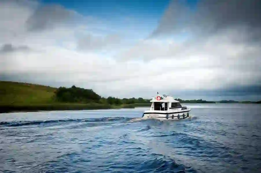 Small white boat sailing across the waters of Lough Erne surrounded by grassy hills and cloudy skies in County Fermanagh.