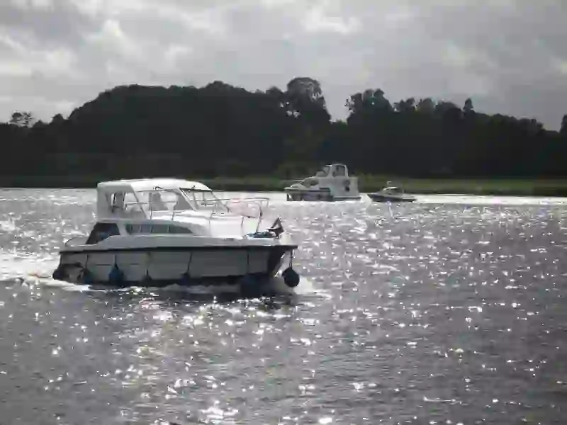 White motorboat cruising across Lough Erne waters with more boats behind and forested hills under cloudy skies.