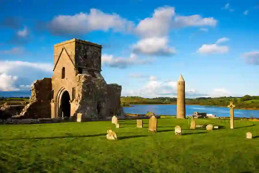 Stone ruins, round tower, and old gravestones at Devenish Monastic Site on Lough Erne with blue sky and water backdrop.