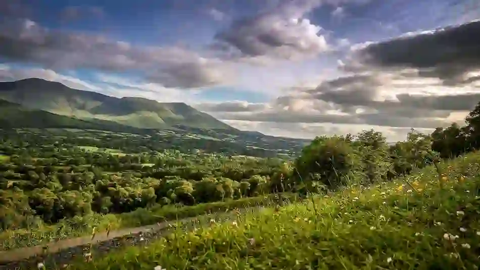 Scenic view of rolling green hills and dramatic mountains under a cloudy sky in Ireland’s Hidden Heartlands.