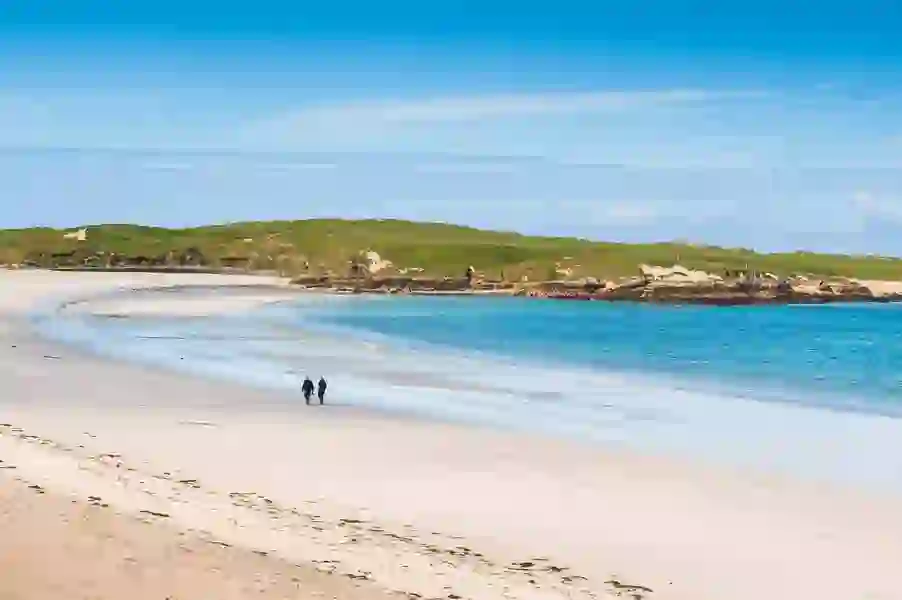 Two people walk along a wide, white sandy beach at Dog's Bay, Galway, with turquoise water and blue sky.