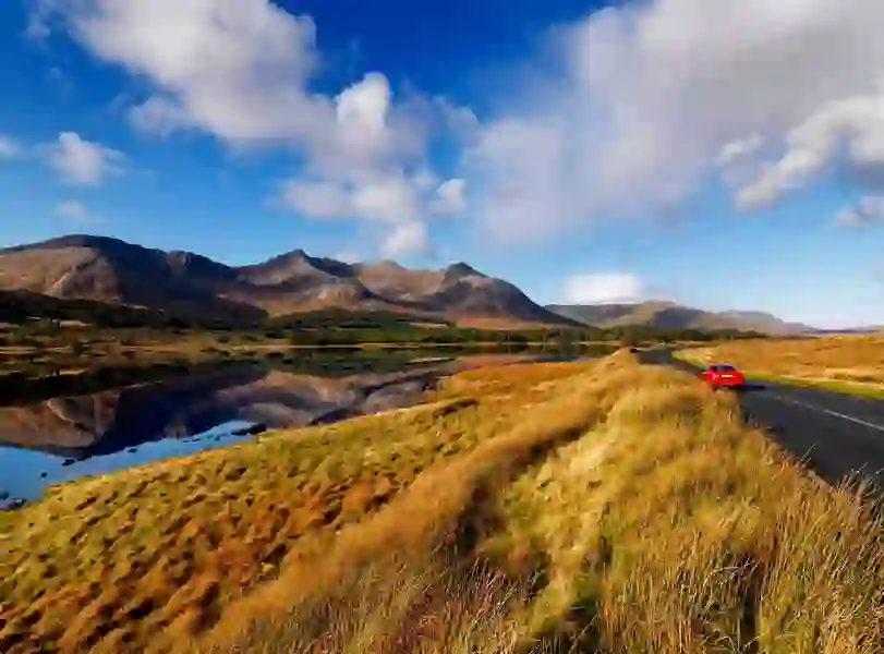 Red car driving past a calm lake with mountain reflections in Connemara under a blue sky.