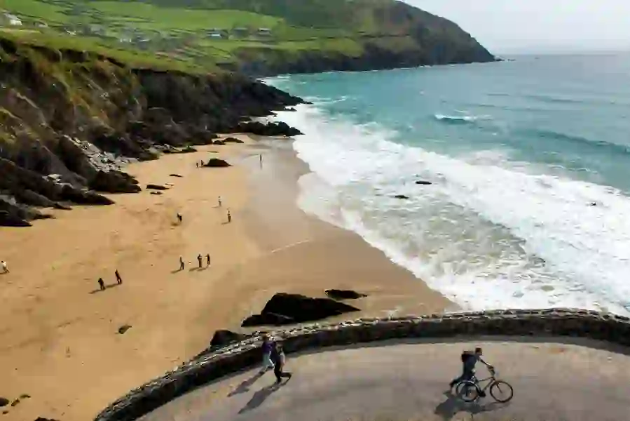 People walking and cycling by sandy Coumeenoole Beach in County Kerry with cliffs and waves.