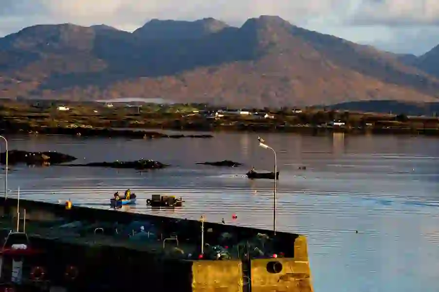 Quiet fishing pier at Roundstone, Galway, with boats and nets, set against calm waters and rugged mountain backdrop.
