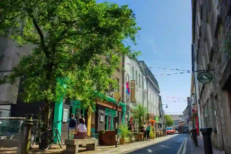 Quiet street in Galway city with bunting, a tree in full leaf, and people outside shopfronts.