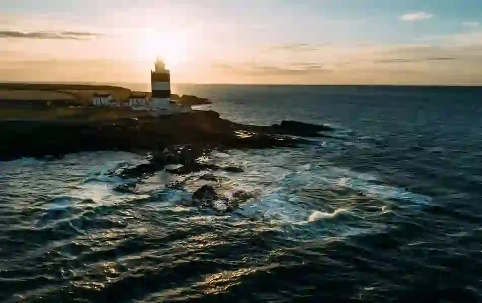 Sunset behind Hook Head Lighthouse in County Wexford, casting a warm glow over the rugged shoreline and Atlantic waves.