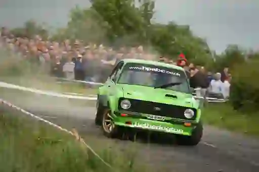 A green Ford rally car drifts through a turn on a wet country road, kicking up spray in front of a crowd.
