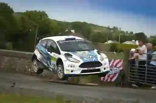 A white rally car airborne over a stone bridge with spectators watching from nearby.