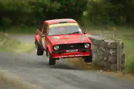 A red Ford rally car mid-jump on a narrow country road flanked by stone walls and greenery.