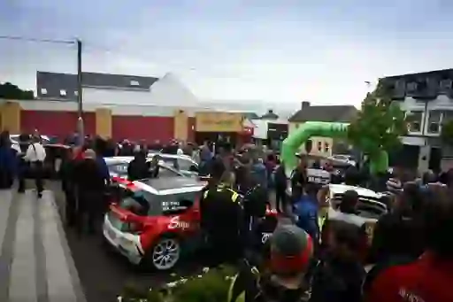 A crowd gathers in a town centre around rally cars at the starting line of the Donegal rally.