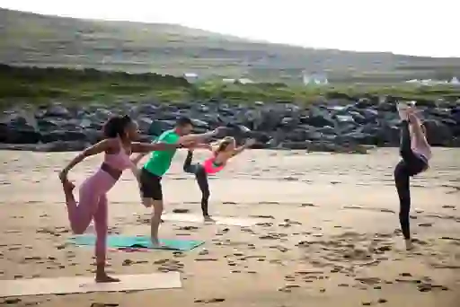 Four people holding balancing yoga poses on mats on Fanore Beach sand with rocky hills behind.