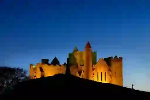 Rock of Cashel illuminated against a deep blue evening sky, with silhouettes in the foreground.