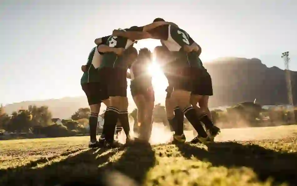 A rugby team in green jerseys forms a huddle at sunset on a grassy pitch, with dramatic backlighting creating a silhouette effect.