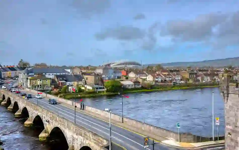A scenic view of Thomond Park stadium in Limerick rising behind the cityscape, with the River Shannon and historic bridge in the foreground.