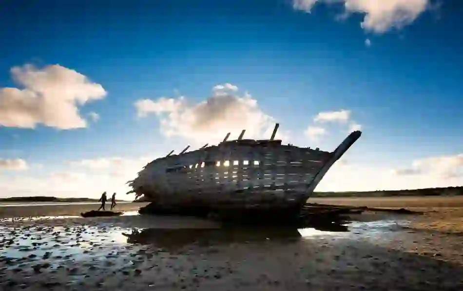 Wooden Bad Eddie shipwreck on Dunbeg beach in County Donegal at sunset with two walkers in silhouette nearby.