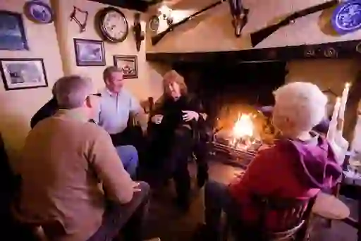 Group of friends chatting around a pub fireplace decorated with clocks and antiques in the Crosskeys Inn, County Antrim.