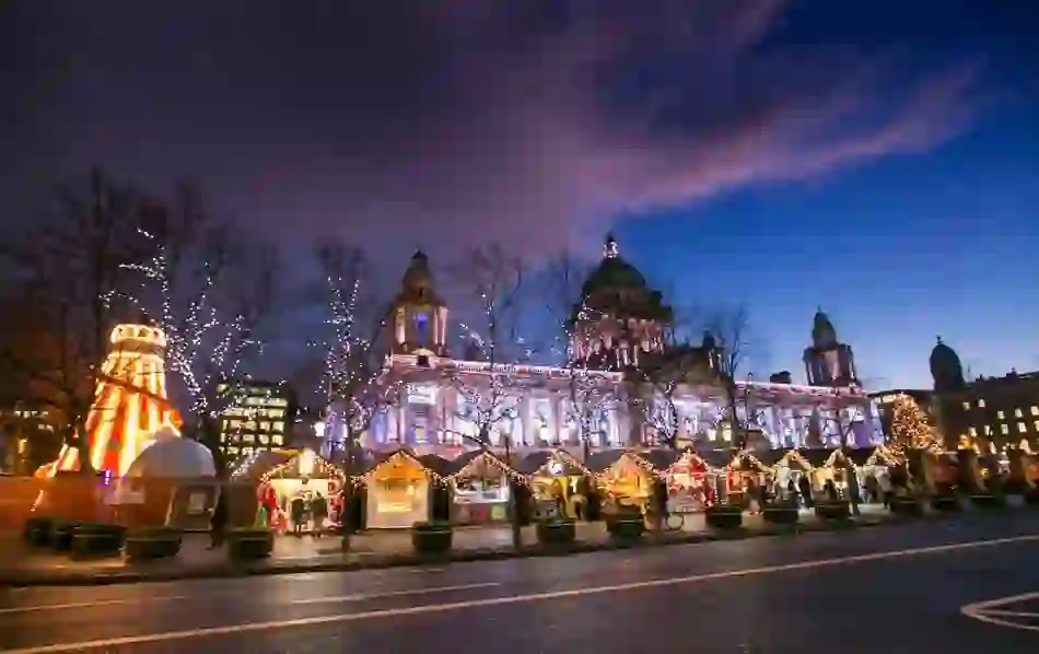 Belfast City Hall sparkles with festive lights above a lively Christmas market at dusk.