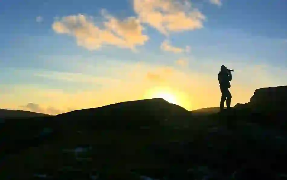 Silhouette of a person with a camera standing on a rocky hilltop in Portacloy, County Mayo, during a golden sunset.
