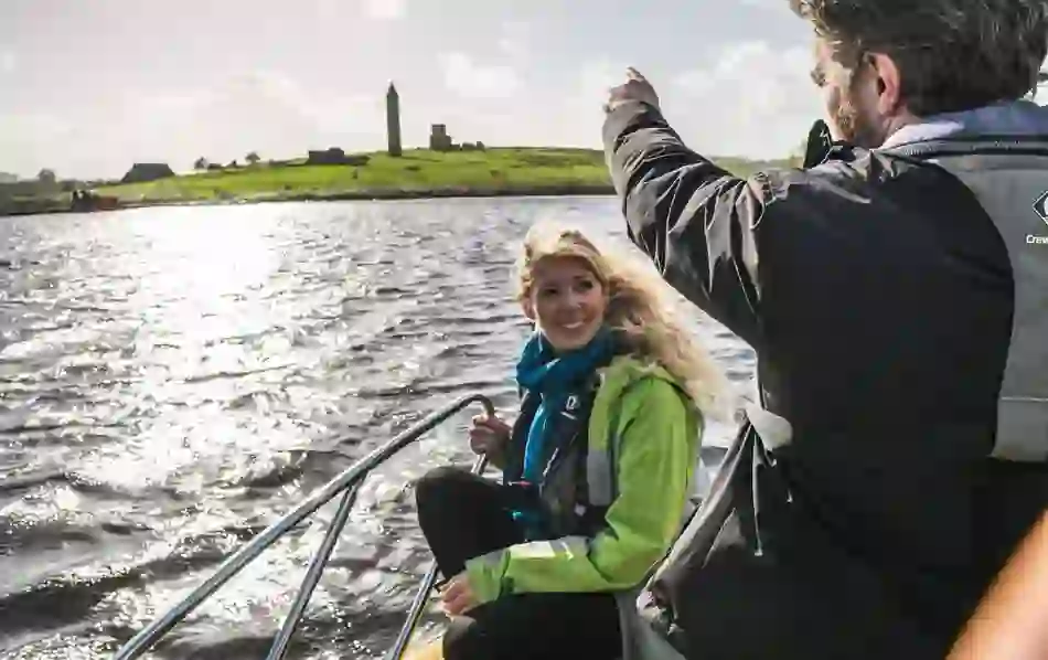 Smiling passenger and guide on a boat taxi across Lough Erne with Devenish Island monastery in the background.