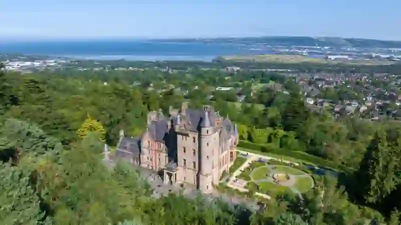 Aerial view of Belfast Castle surrounded by woodland with Belfast Lough and the city skyline stretching out in the distance below.