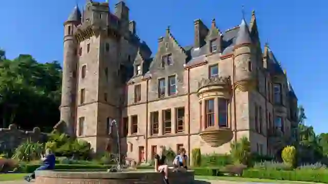 Belfast Castle’s stone turrets overlooking landscaped gardens, with visitors relaxing by a fountain on a sunny day.