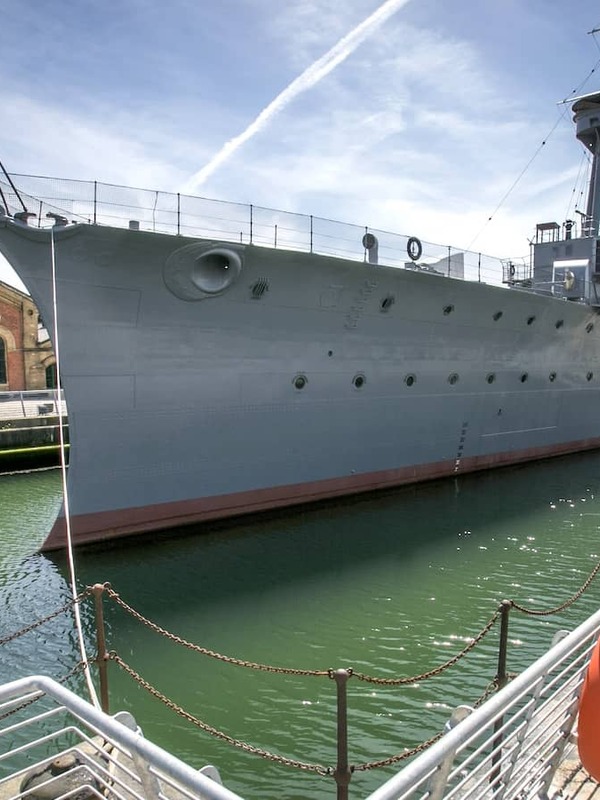HMS Caroline moored at Titanic Quarter, Belfast, with historic brick buildings and harbour walkway.