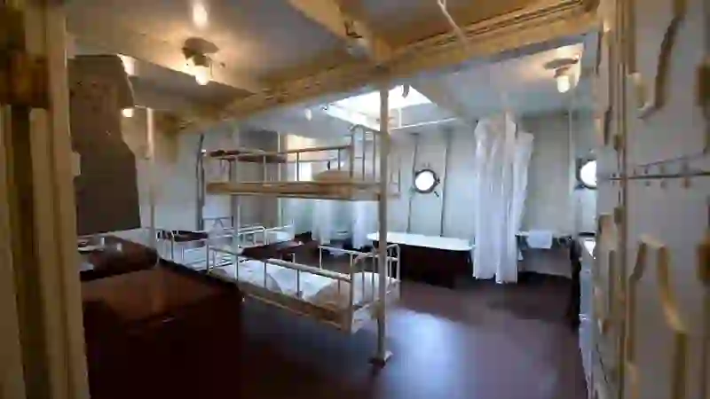 Sleeping quarters on HMS Caroline with metal bunk beds, curtained wash area and porthole windows.