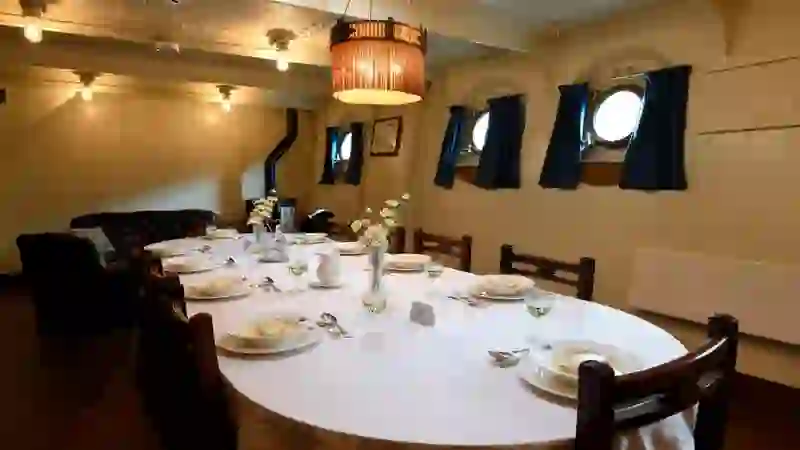 Dining room set for a meal on HMS Caroline with white tablecloths, place settings and porthole windows.