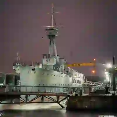 HMS Caroline moored in Belfast's Titanic Quarter at night with Harland & Wolff cranes in the background.