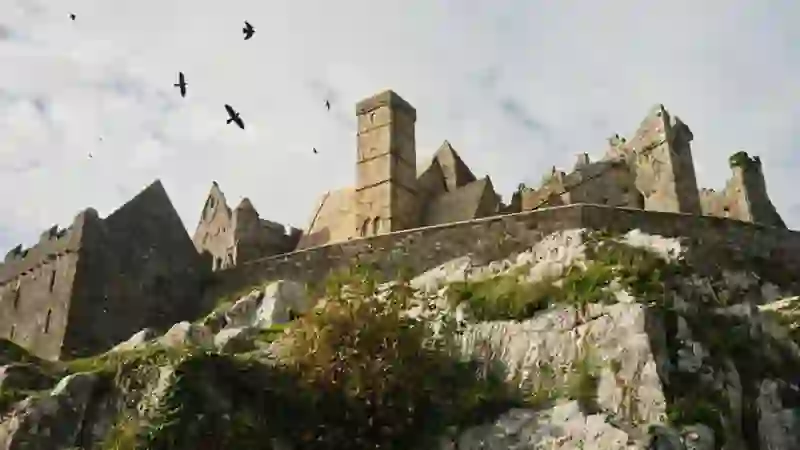 Rock of Cashel, County Tipperary, perched on limestone cliffs under a cloudy sky with birds overhead.