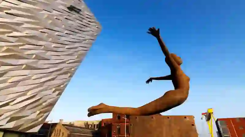Rust sculpture of a diving figure beside the angular façade of Titanic Belfast under a clear blue sky.