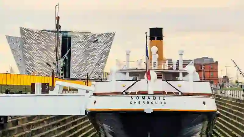 SS Nomadic moored in dry dock with the modern Titanic Belfast building rising behind it.