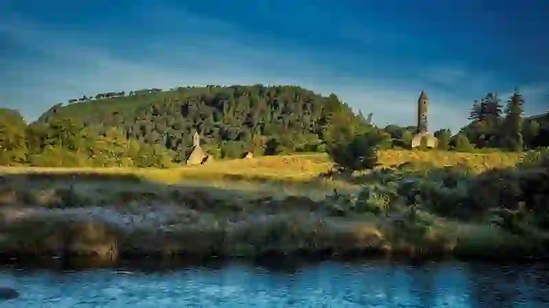 Round tower and stone church beside meadow and river at Glendalough in Wicklow Mountains National Park.