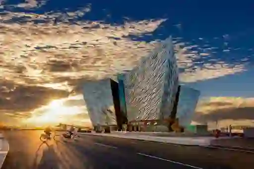 Two cyclists heading towards Titanic Belfast as the sun rises against a blue sky with some clouds.