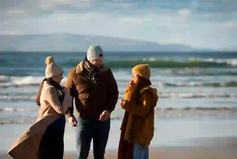 Friends wrapped in winter layers chatting and walking along a breezy Portrush Strand, County Antrim, with waves behind them.