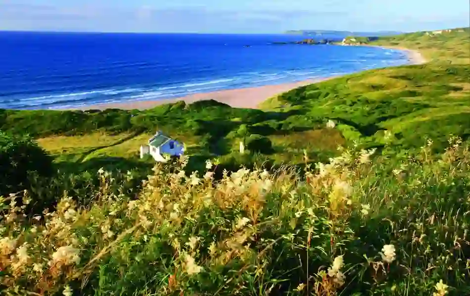 A small cottage overlooks golden sand and blue Atlantic surf along a lush green coastline in summer in Donegal.