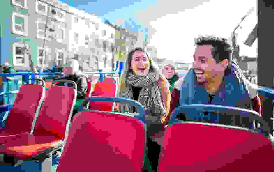 Smiling passengers on the upper deck of an open-top sightseeing bus on a bright day in Cork city.