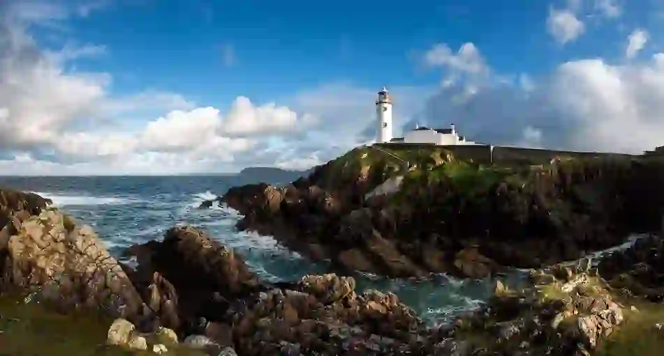 White lighthouse at Fanad Head stands above crashing waves and rugged cliffs under a bright blue sky.